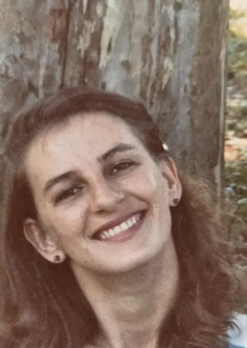 Retrato de Zeli jovem, sorrindo, de blusa listrada, diante de uma árvore.