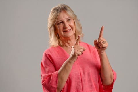 Dra. Zeli, de blusa coral, aponta para cima com um sorriso, em retrato de estúdio.